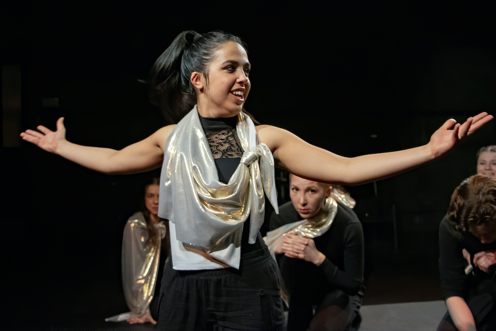 Actress standing with arms outstretched in mid-performance while other actors look on.